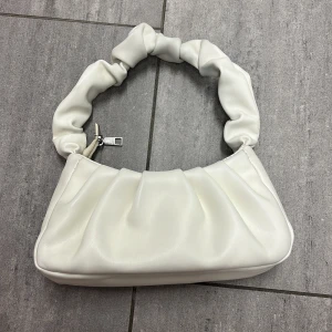 Vit axelväska med scrunchie-band - Säljer en superfin vit axelväska med scrunchie-inspirerat handtag. Väskan har en mjuk, puffig look och stängs med dragkedja. Perfekt storlek för det viktigaste och ger en trendig touch till din outfit.