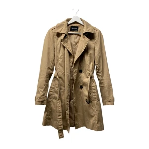 Beige trenchcoat från Stradivarius M - Snygg beige trenchcoat från Stradivarius i storlek M. Jackan har klassisk dubbelknäppning, bälte i midjan och långa ärmar. Tillverkad i 100% polyester, perfekt för vår och höst. Stilsäker och tidlös modell med snygga detaljer.