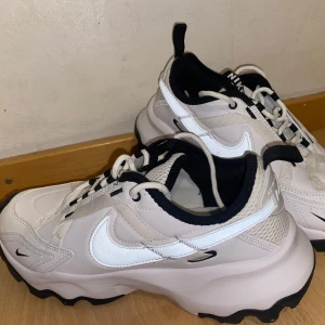 Chunky vita sneakers från Nike - Säljer ett par chunky vita Nike sneakers med svart sula och svarta detaljer. Skorna har en vågig plattformssula, stora vita swoosh-loggor på sidorna och meshpartier för extra andningsförmåga. Perfekta för dig som gillar streetstyle och vill sticka ut. Oanvända. Storlek 41
