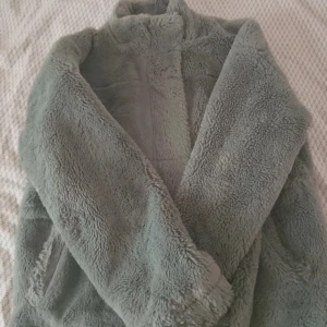 Grå teddyjacka med hög krage - Supermjuk och fluffig grå teddyjacka med hög krage och dragkedja framtill. Perfekt för kyliga dagar när du vill vara både varm och snygg. Jackan har en loose passform och är tillverkad i gosig syntetpäls.