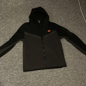 Svart Nike hoodiejacka med dragkedja - Svart hoodiejacka från Nike med hel dragkedja och huva. Jackan har en orange Nike-logga på bröstet och diskreta detaljer längs bröstet och ärmarna. Perfekt för chill dagar eller när du vill ha en sportig look.