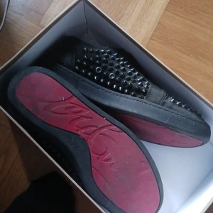Christian Louboutin med nitar - Svarta sneakers från Christian Louboutin med ikoniska röda sulor och coola svarta nitar. Skorna är perfekta för dig som vill ha något extra edgy i din outfit.