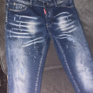 Dsquared2 ICON blå jeans med slitningar - Säljer ett par blå Dsquared2 ICON jeans med coola slitningar och färgstänk på framsidan. Jeansen har en snygg tvättad look, klassiska fem fickor och en stor vit patch med röd text bak i midjan. Perfekt för dig som gillar streetstyle och vill sticka ut.