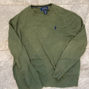 Olivgrön tröja från Polo Ralph Lauren - Olivgrön tröja från Polo Ralph Lauren i mjuk bomull. Klassisk rund halsringning och diskret broderad logga i mörkblått på bröstet. Långärmad modell med ribbade muddar och nederkant. Perfekt för en chill och stilren look.