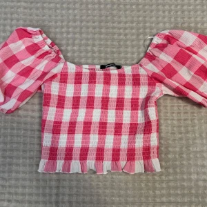 Rutig rosa topp från Gina Tricot - Supersöt rutig topp från Gina Tricot i rosa och vitt. Modellen är croppad med volangkant nertill och har puffiga korta ärmar som sitter offshoulder. Endast använd en gång. Storlek s