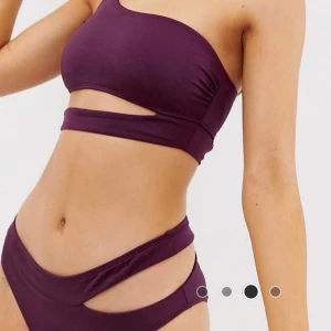 Lila asymmetrisk bikini med cut-outs - Snygg bikini i mörklila färg. Toppen har en axel och cool cut-out detalj under bysten. Bikinitrosan har matchande cut-outs på sidorna för en trendig look. Tillverkad i stretchigt syntetmaterial som sitter skönt på kroppen. Perfekt för dig som vill sticka ut på stranden! ÖVERDELEN ÄR I STORLEK EU 44 OCH UNDERDELEN I STORLEK EU 38