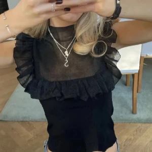 Volang topp - Supersnygg svart blus med transparent mesh upptill och volangdetaljer längs axlar och bröst. Hög hals och ärmlös design ger en trendig look. Perfekt att styla med jeans eller kjol för en cool outfit.