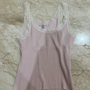 Rosa ribbad linnetopp med spets - Supersöt ljusrosa ribbad linnetopp från Brandy Melville med tunna vita spetsdetaljer längs urringning och axelband. Perfekt för lager på lager eller sommarens varma dagar. Smal passform och diskret randigt mönster.