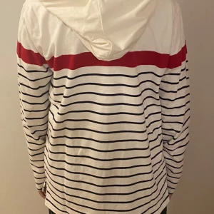 Randig hoodie från Hope & Henry XL - Vit hoodie med marinblå ränder och en bred röd rand över bröstet från Hope & Henry. Tröjan har huva, knappar vid halsen med blå detalj och är långärmad. Tillverkad i mjuk bomull, perfekt för en avslappnad stil.