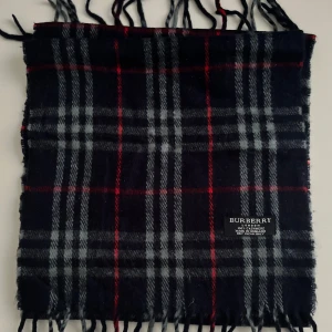 Rutig ullhalsduk från Burberry - Klassisk halsduk från Burberry i svart, grått och rött rutmönster. Gjord i 100% ull med fransar i kanterna. Perfekt accessoar för att lyfta din outfit med en tidlös och stilren look.