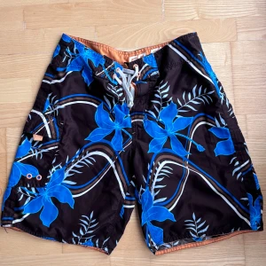  badshorts från Body Zone - Snygga badshorts från Body Zone med svart bas och stora blå blommor samt bruna och vita vågiga linjer. Shortsen har snörning i midjan, meshfoder och praktisk ficka på sidan. Perfekta för stranden eller poolen!