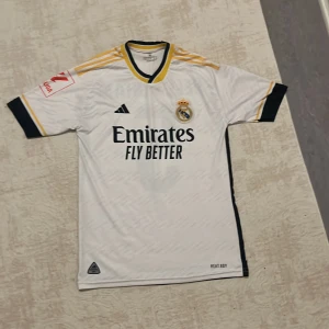 Real Madrid Bellingham 5 Adidas tröja M - Real Madrid 2024 matchtröja med Bellingham 5 på ryggen. Vit med svarta och gula detaljer, Adidas-logga och klubbmärke på bröstet. Kortärmad, tillverkad i lätt och ventilerande AEROREADY-material. Snygga detaljer på ärmar och axlar.
