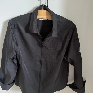 C.P. Company overshirt  - Svart overshirt från C.P. Company med klassisk krage, dragkedja framtill och ikonisk linsdetalj på ärmen. Den är i nyskick. Är L men sitter som M.
