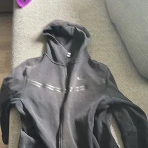 Svart Nike vindjacka med huva M - Svart vindjacka från Nike med huva och hel dragkedja. Jackan har en diskret logga på bröstet, reflekterande detalj över bröstet och en praktisk ficka med dragkedja på ärmen. Perfekt för dig som gillar sportig och clean stil. Från Jd Sports