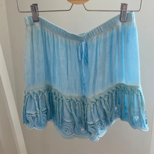 Ljusblå kortkjol med volang och spets - Supersöt ljusblå kortkjol med volangkant och broderad spets nertill. Kjolen har knytband i midjan och coola flätade detaljer längs kanten. Väldigt somrig och luftig! Knappt använd.