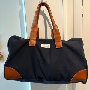 Weekend väska  - Snygg och rymlig mörkblå axelväska / weekend bag från lager 157 med bruna handtag och hörn i skinnimitation. Väskan har dragkedja upptill och en stilren etikett framtill. Perfekt för dig som vill ha en stilren och praktisk väska till vardagen. Allt är i fint skick förutom att dragkedjan inte går att dra lås