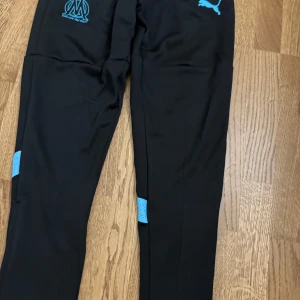 Svarta träningsbyxor Puma Marseille - Svarta träningsbyxor från Puma med Olympique de Marseille-logga i turkosblått på låret. Byxorna har smal passform, elastiskt material och ventilerande detaljer i blått vid knäna. Perfekta för fotboll eller träning.