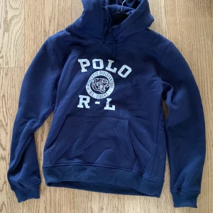 Marinblå hoodie Polo Ralph Lauren - Snygg marinblå hoodie från Polo Ralph Lauren med vit tryckt logga och tiger på bröstet. Klassisk känguruficka, huva med snörning och mjukt bomullsmaterial. Storlek L skulle säga att den är ungefär som en M. Har använts ett fåtal gånger. Skriv gärna om det är något du undrar! Pris kan diskuteras vid snabb affär!