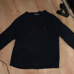 Mörkblå kabelstickad tröja Polo Ralph Lauren - Kabelstickad svart tröja från Polo Ralph Lauren med klassisk röd broderad logga på bröstet. Tröjan har rund halsringning, ribbade muddar och är långärmad. Perfekt för dig som gillar stilren och tidlös design.