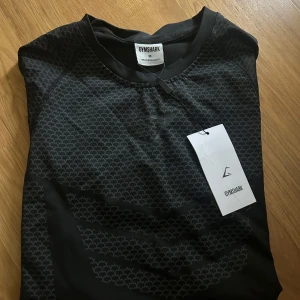 Svart mönstrad t-shirt från Gymshark - Snygg svart t-shirt från Gymshark med diskret geometriskt mönster över hela tyget. T-shirten har rund halsringning och är tillverkad i ett lätt och stretchigt material som passar perfekt för träning. Stilren logga på bröstet och modern passform.