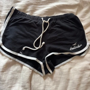 Svarta high-rise shorts Hollister - Svarta high-rise shorts från Hollister med vit snörning i midjan och vita kanter. Mjukt bomullsmaterial och sportig look, perfekt för sommardagar. Broderad Hollister-logga framtill. Snygg och enkel design med bekväm passform.