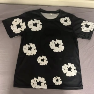 Svart denim tears T-shirt  - Cool svart t-shirt med stora vita blommiga mönster över hela tröjan. Klassisk rund halsringning och korta ärmar. Perfekt för dig som vill sticka ut med en unik och trendig stil.