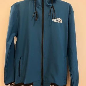 Blå vindjacka från Sport Life, strl L - Blå vindjacka med huva från Sport Life. Jackan har svarta detaljer, dragsko i huvan och dragkedja framtill. Elastisk nederkant med The North Face-tryck runt om. Perfekt för dig som gillar en sportig och clean stil.