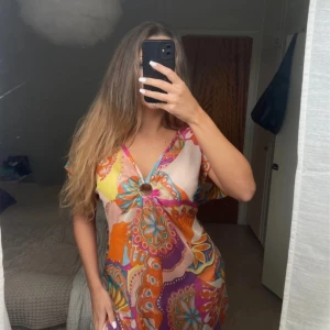 Färgglad kortklänning med ringdetalj - Säljer en superfin kortklänning i färgglatt mönster med orange, lila, rosa, gul och blå toner. Klänningen har v-ringning, korta ärmar och en snygg ringdetalj framtill. Perfekt för sommarens alla tillfällen och ger riktiga retrovibbar. Lite paljett detaljer. Använd en gång 