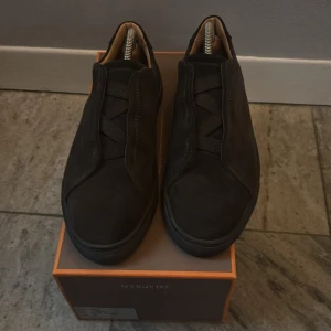 Mörkbruna sneakers från Myrqvist - Snygga mörkbruna sneakers från Myrqvist i mocka med elastiska band istället för klassisk snörning. Skorna har en stilren design med rund tå och svart platt sula. Insidan är fodrad i ljust skinn för extra komfort. Perfekta för en clean och modern look.
