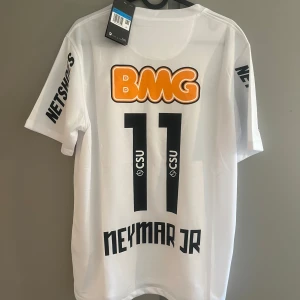 Santos Neymar Jr #11 Nike fotbollströja - Vit Santos fotbollströja med Neymar Jr och nummer 11 på ryggen. Tröjan har tryckta sponsorer som BMG och Netshoes, Santos klubbmärke på bröstet och Nike-logga. Tillverkad i lätt Dri-FIT-material som andas, perfekt för match eller träning.