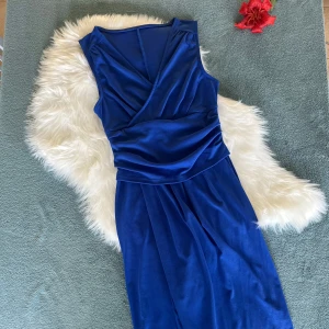 Blå omlottklänning i satinlook - Supersnygg blå långklänning med omlottliv och ärmlös design. Klänningen har en elegant v-ringning och är gjord i ett glansigt material som ger en lyxig känsla. Perfekt för dig som vill sticka ut med en klassisk men modern look.