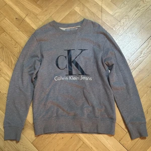 Grå sweatshirt från Calvin Klein Jeans - Snygg grå sweatshirt från Calvin Klein Jeans med stor svart CK-logga och vit text framtill. Klassisk rund halsringning och långa ärmar. Perfekt för en avslappnad och stilren look. Tillverkad i mjuk bomullsmix för extra komfort.