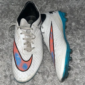 Nike Hypervenom fotbollsskor - Nike Hypervenom fotbollsskor i vitt med blå och orange swoosh-detaljer. Skorna har snörning, mönstrad ovandel och blå sula med dobbar för gräsplan. Perfekta för dig som vill sticka ut på planen med snygg design och bra grepp.