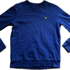 Blå Lyle & Scott sweatshirt i bomull - Snygg blå sweatshirt från Lyle & Scott med klassisk rund hals och gul broderad logga på bröstet. Tröjan är i mjuk bomull och har ribbade muddar vid ärmslut och nederkant. Perfekt för dig som gillar stilren och enkel design.