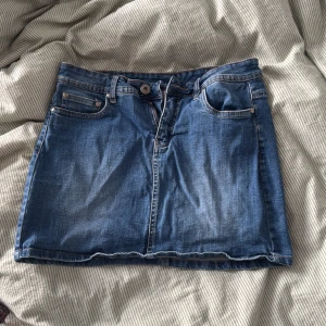 Blå jeanskjol från Millie, storlek 36 - Snygg blå jeanskjol från Millie i klassisk modell med fem fickor, bälteshällor och knapp- samt dragkedjestängning. 