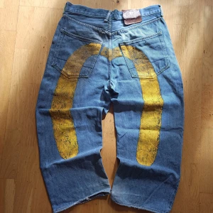 Evisu baggy jeans - Unika baggy jeans från Evisu i blå denim med handmålat gult måsmotiv på baksidan. Klassisk femficksmodell, raka och vida ben med croppad längd. 100% bomull och coola kontrastsömmar. Perfekta för dig som vill sticka ut med streetwear-vibe. För mer information, bilder och prisdiskutering i dm. Äkta