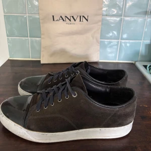 Svarta sneakers från Lanvin med lackad tå - Snygga svarta sneakers från Lanvin med en cool lackad tå och mocka på sidorna. Skorna har svarta snören och vit platt sula med Lanvin-logga undertill. Perfekt för dig som gillar stilrena och lyxiga detaljer i din vardagslook. Köpta från Plick storlek 45,5 46