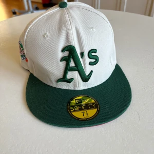 Oakland Athletics 59FIFTY keps vit/grön - Snygg 59FIFTY keps från New Era med Oakland Athletics A's broderat i grönt framtill. Kepsen är vit med grön skärm och har broderad 1989 World Series patch på sidan. Undersidan av skärmen är röd och insidan har klassisk 59FIFTY branding.