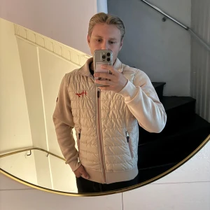 Beige cardigan från Hugo - Snygg beige cardigan från Hugo med quiltad framsida och broderad röd logga på bröstet. Jackan har dragkedja framtill, två sidofickor med dragkedja och mjuka ärmar i bomullsmix. Perfekt för dig som gillar stilren design med en twist. Priset går att diskutera, passar M/L
