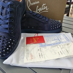 Mörkblå Christian Louboutin sneakers - Snygga mörkblå sneakers från Christian Louboutin i mocka med ikoniska röda sulor och coola svarta nitar på tån. Skorna har rund tå, snörning och lyxig känsla. Perfekta för dig som vill sticka ut med exklusiv stil.