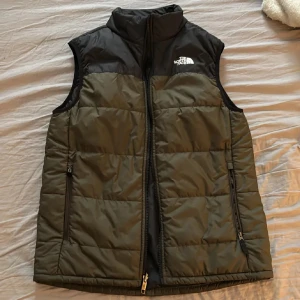 Svart och olivgrön dunväst The North Face - Säljer en dunväst från The North Face i svart och olivgrönt. Västen har hög krage, två dragkedjefickor och logga på bröstet. Perfekt för lager-på-lager och outdoor-vibe. Stängs med dragkedja hela vägen och har quiltad design. Barnstorlek så passar xs/s