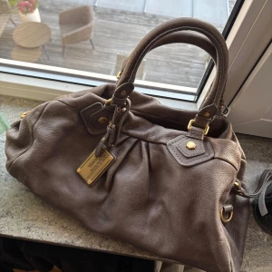 Marc by Marc Jacobs brun handväska skinn - Säljer en brun handväska i äkta skinn från Marc by Marc Jacobs. Väskan har guldfärgade metalldetaljer, dubbla handtag och avtagbar axelrem. Insidan är fodrad med svartvitt mönster med bokstäver