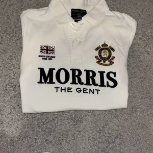 Vit Morris Gents piké XS - Vit pikétröja från Morris Gents i bomull, med broderade detaljer och tryck. Stor svart logga på bröstet, emblem och brittisk flagga, samt siffran 68 på ärmen. Klassisk krage med knappar och snygg passform. Perfekt för dig som gillar stilrena och sportiga plagg.