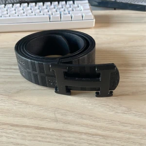 Svart Bälte med H-spänne - Snyggt svart bälte med klassiskt H-spänne i metall. Bältet har ett diskret rutigt mönster och är gjort i skinn. Perfekt accessoar för att lyfta din outfit med en lyxig touch.