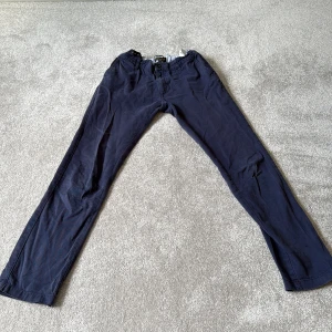 Mörkblå chinos från Lyle & Scott - Snygga mörkblå chinos från Lyle & Scott med klassisk gul logga broderad på bakfickan. Byxorna har rak passform, knappstängning och elastisk midja för extra komfort. Tillverkade i mjuk bomull, perfekta för en chill och stilren look.