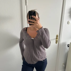 Lila långärmad topp Odd Molly spets - Supersöt lila långärmad topp från Odd Molly med ribbad struktur och spetsdetaljer i v-ringningen. Liten rosett framtill och vågig kant nertill ger en extra gullig touch. 