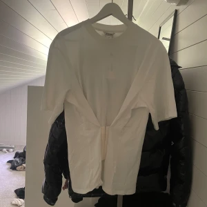 Vit t-shirt med cool cutout från Monki - Vit monki t-shirt med korsett knäppe i midjan. Jätte snygg till jeans och kostym byxor för en avslappnad men prydlig look! Helt ny endast testad och har en liten smink fläck som går bort i tvätten!🥰