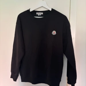 Svart sweatshirt från Moncler - Svart sweatshirt från Moncler med rund hals och lång ärm. Tröjan har ett broderat Moncler-märke på bröstet.