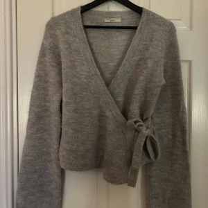 Grå omlottkofta från Urban Outfitters - Hej , jag säljer en mysig grå omlottkofta med knyt i sidan och v-ringad hals. Koftan är stickad och har långa ärmar samt ribbade muddar. Perfekt för lager-på-lager och chill dagar. Passar dig som gillar enkel och stilren look. Super söt kofta💕