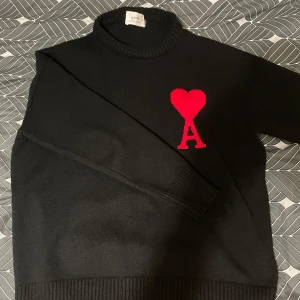 Svart stickad tröja från Ami Paris - Svart stickad tröja från Ami Paris med röd hjärtlogga och bokstaven A på bröstet. Tröjan har ribbade muddar och hög hals, perfekt för dig som gillar stilrena plagg med en twist. Klassisk passform och långärmad modell.